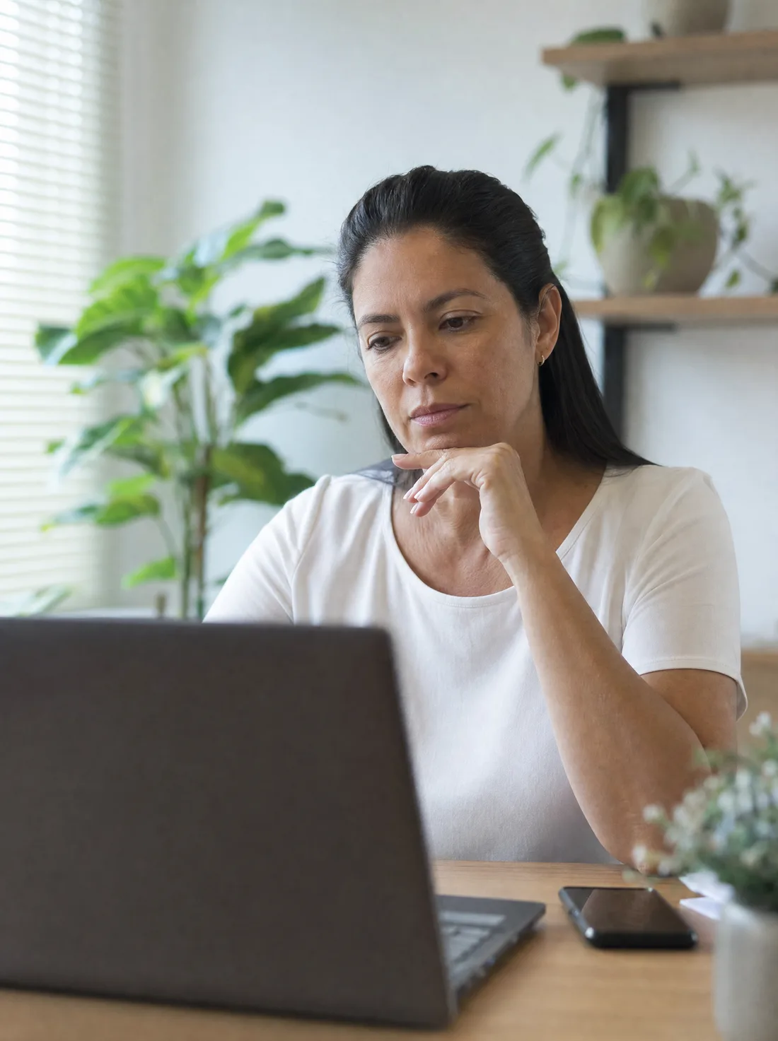Profissional sentada à mesa do consultório, apoiada na mão, avaliando a tela do notebook com o celular ao lado